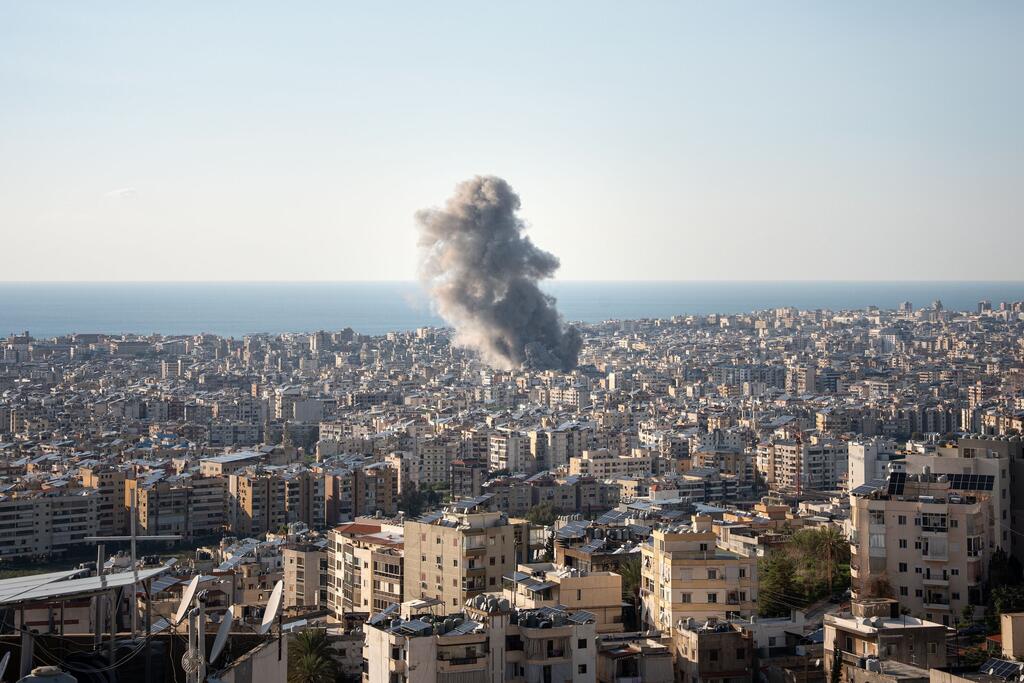 Aerial view of a densely populated city in Lebanon with a large plume of dark smoke rising from an explosion in the distance, with the sea visible on the horizon.