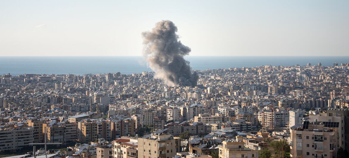 Aerial view of a densely populated city in Lebanon with a large plume of dark smoke rising from an explosion in the distance, with the sea visible on the horizon.