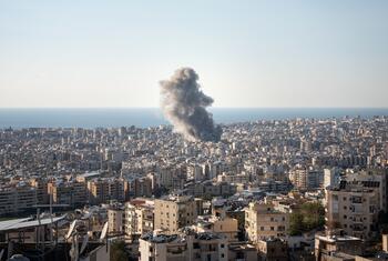 Aerial view of a densely populated city in Lebanon with a large plume of dark smoke rising from an explosion in the distance, with the sea visible on the horizon.