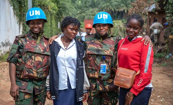 Wanawake wawili wa Tanzania wanaofanya kazi ya kulinda amani ya Umoja wa Mataifa wakiwa wamevaa mavazi ya kujificha pamoja na kofia za UN za bluu wamesimama pamoja na wanawake wawili wa eneo hilo katika mazingira ya vijijini.