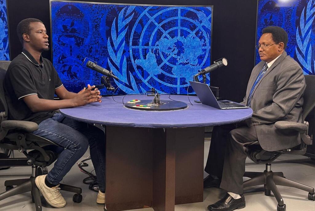 Wanaume wawili wanakaa kwenye meza ya mviringo wakiwa na maikrofoni katika studio wakati wa mahojiano, wakiwa na nembo kubwa ya Umoja wa Mataifa inayoonekana kwenye skrini nyuma yao.