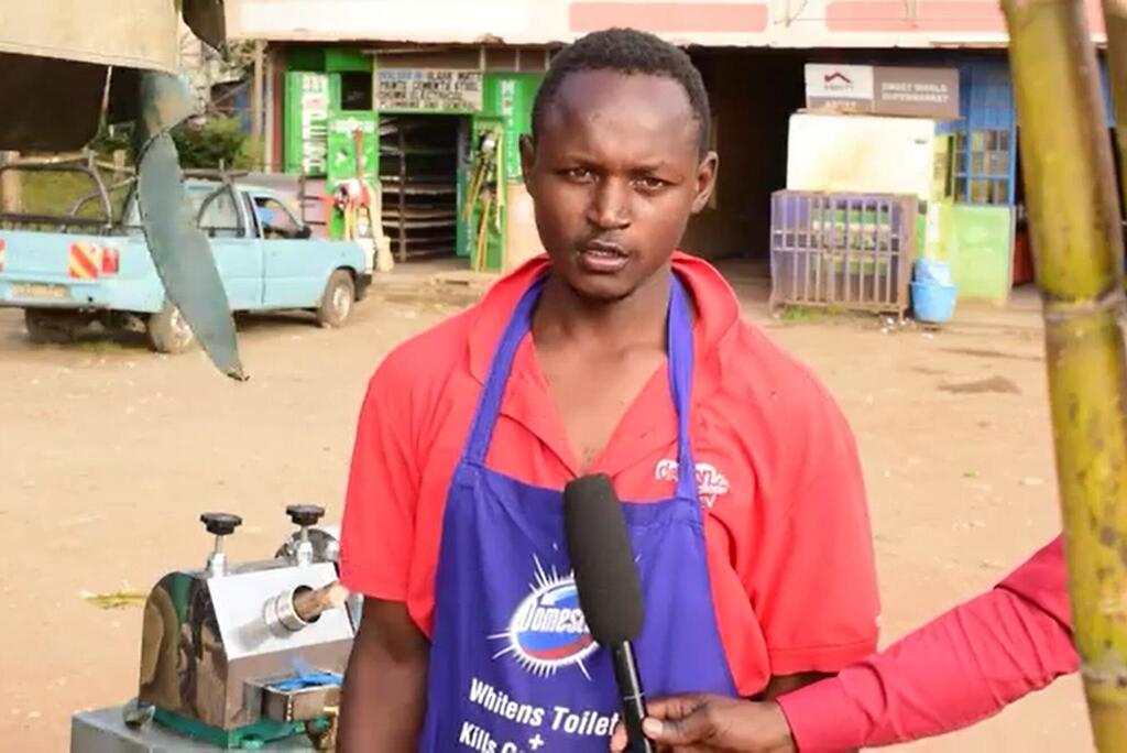 Kijana mweusi mwenye shati nyekundu na aproni ya bluu anahojiwa barabarani huko Kajiado, Kenya, akiwa amesimama kando ya mashine ya kutoa juisi ya miwa.