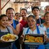 Um grupo de cozinheiros timorenses sorridentes, usando aventais, posa em uma cozinha segurando pratos preparados.