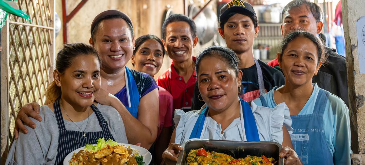 Um grupo de cozinheiros timorenses sorridentes, usando aventais, posa em uma cozinha segurando pratos preparados.