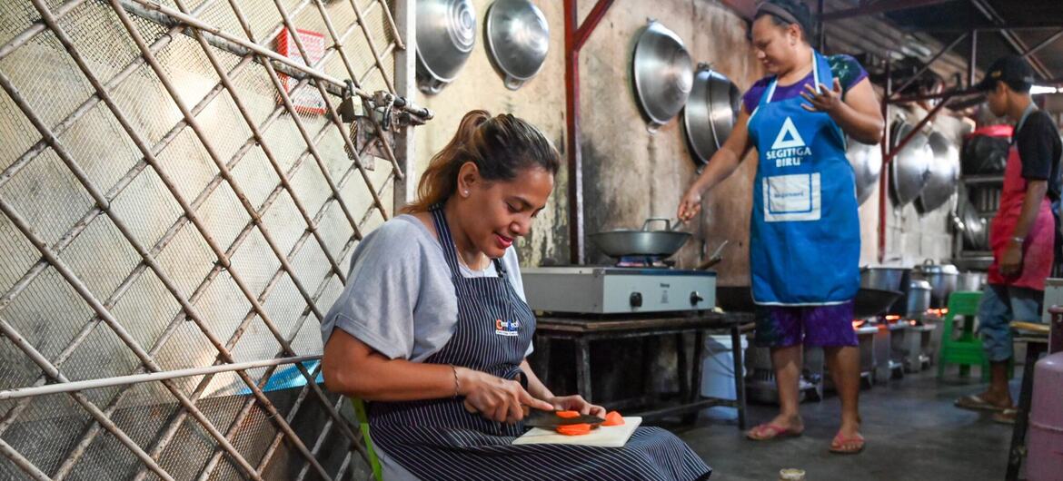 Uma mulher com um avental listrado senta-se na cozinha cortando legumes enquanto outra pessoa cozinha ao fundo.