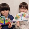 Dois meninos estão lado a lado em uma sala de aula, cada um segurando um prato de papel colorido decorado com penas e tinta.