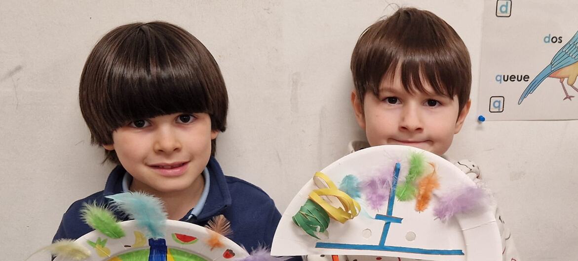 Dois meninos estão lado a lado em uma sala de aula, cada um segurando um prato de papel colorido decorado com penas e tinta.