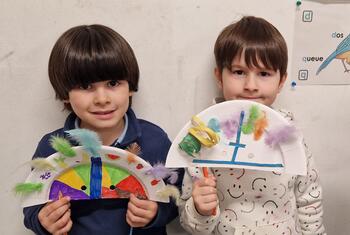 Dois meninos estão lado a lado em uma sala de aula, cada um segurando um prato de papel colorido decorado com penas e tinta.