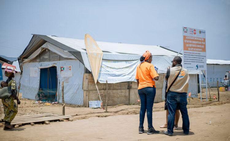 Mwanamke anayebeba mtoto anapita karibu na hema la muda la matibabu katika kambi ya wakimbizi ya Musenyi nchini Burundi, huku wafanyakazi wa UNFPA wakiwa wamesimama karibu.