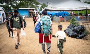 A migrant family, including a woman with a large backpack and a small child, walks through a temporary shelter area at the Lajas Blancas reception centre in Darien, receiving aid from IOM staff.