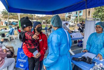 A 5-year-old Venezuelan refugee boy, Deivid, is held by his mother Enyeli during a UNHCR community health event in Lima, Peru. They are receiving vaccinations and medical care alongside other refugee families.