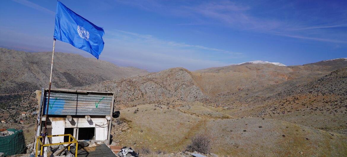 A UNIFIL peacekeeper outpost with a blue UN flag flying atop a small structure, situated in a rugged mountainous landscape in Lebanon near the Blue Line.