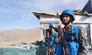 Two UNIFIL peacekeepers in blue helmets and vests stand guard near a damaged structure, overseeing the Blue Line in Lebanon.