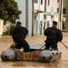 Dois agentes de resgate portugueses navegam numa rua inundada num pequeno barco inflável, remando através de águas lamacentas perto de edifícios residenciais.