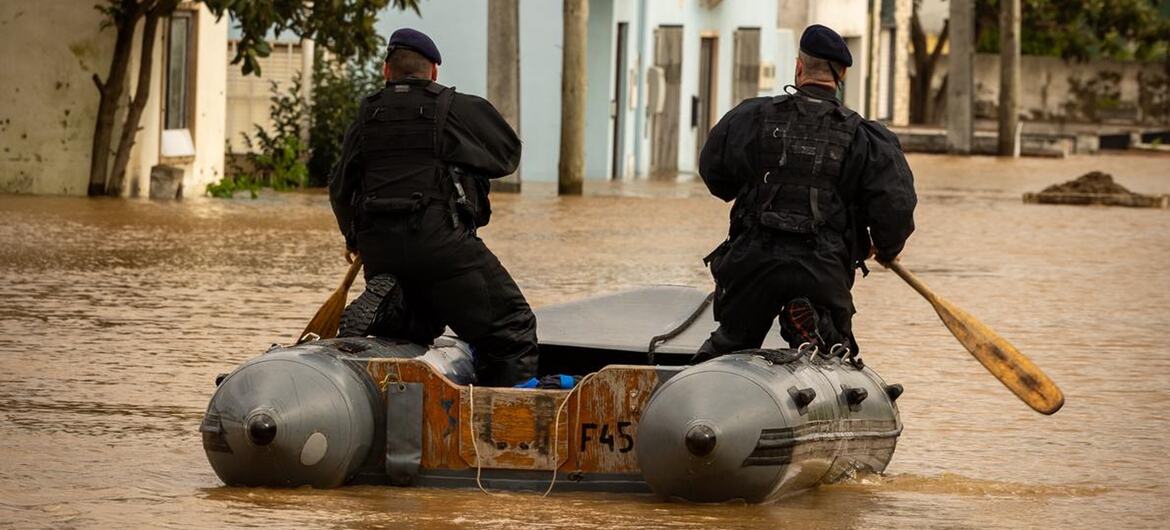 Dois agentes de resgate portugueses navegam numa rua inundada num pequeno barco inflável, remando através de águas lamacentas perto de edifícios residenciais.
