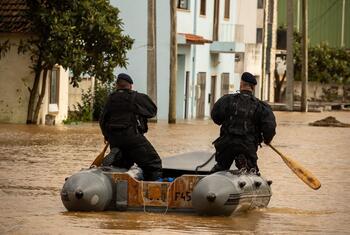 Dois agentes de resgate portugueses navegam numa rua inundada num pequeno barco inflável, remando através de águas lamacentas perto de edifícios residenciais.
