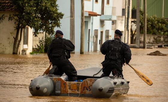 Dois agentes de resgate portugueses navegam numa rua inundada num pequeno barco inflável, remando através de águas lamacentas perto de edifícios residenciais.