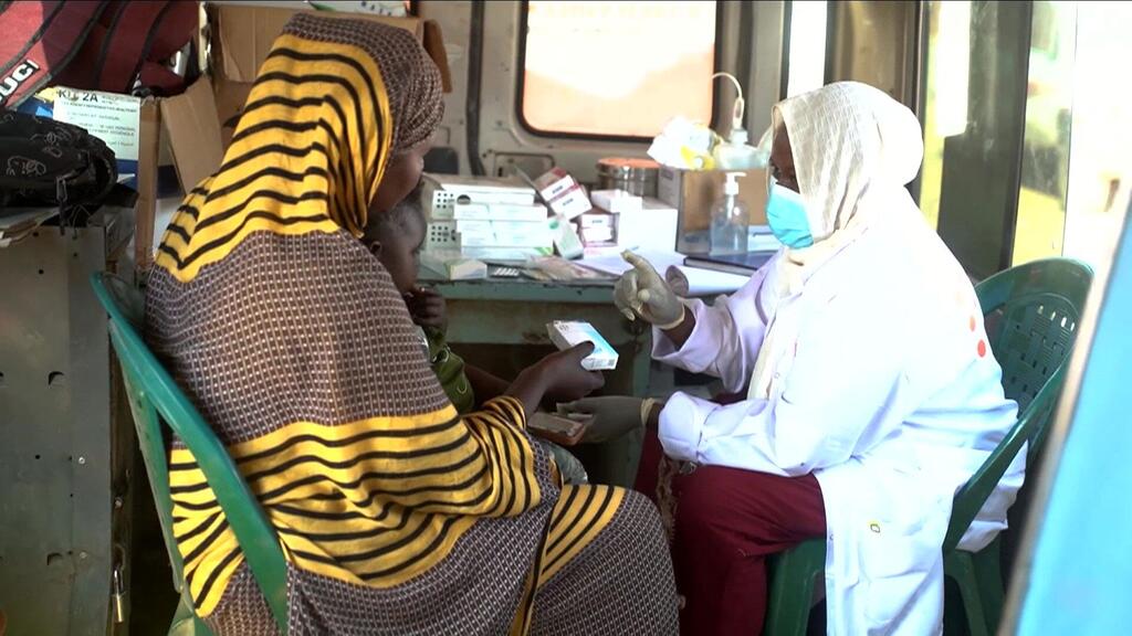 Mfanyakazi wa huduma ya afya mwenye kanzu nyeupe na kinyago anamsaidia mwanamke aliyevaa hijabu yenye mistari katika kliniki ya muda katika kambi ya Abu Al Naja, Sudan, akitoa huduma ya mama wakati wa mgogoro wa kibinadamu unaoendelea.