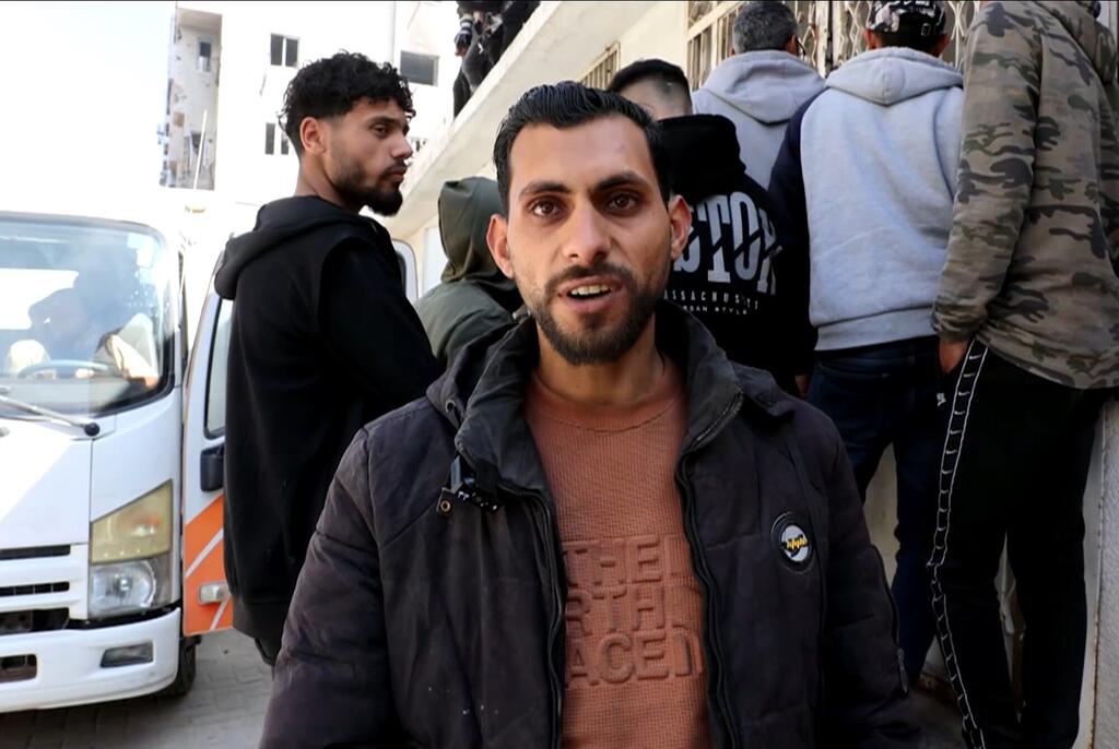 Mwanamume aliyevaa koti nyeusi anasimama mbele ya lori nyeupe, akiwa amezungukwa na watu wengine katika eneo la mijini.