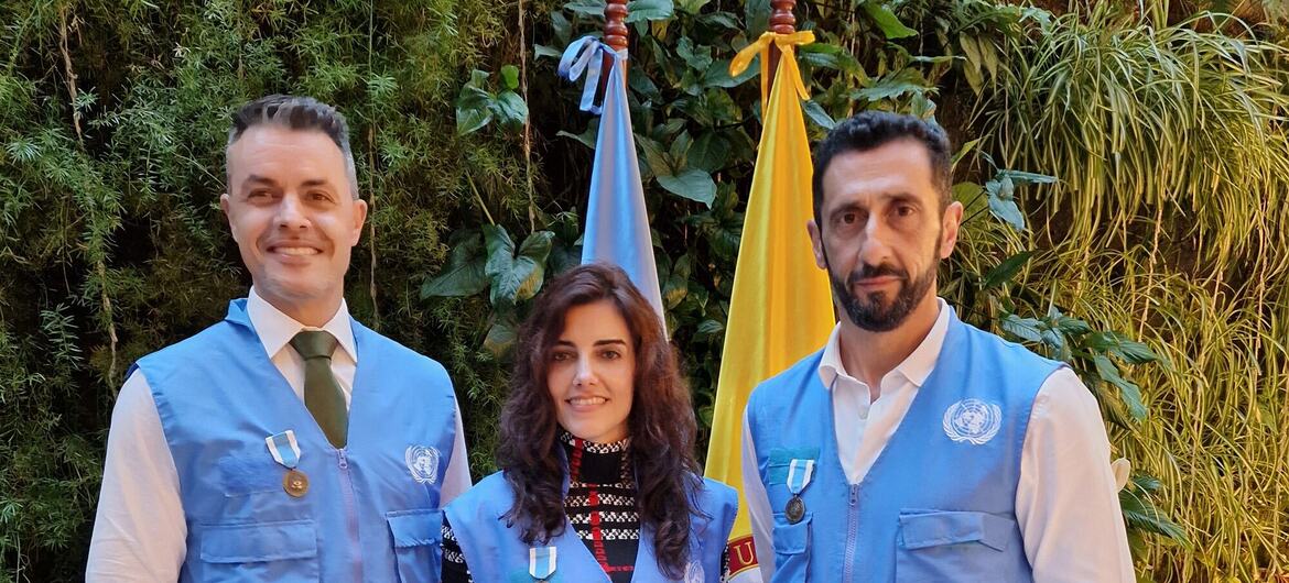 Um retrato de três peacekeepers da ONU, dois homens e uma mulher, vestindo coletes azuis com medalhas, de pé na frente de bandeiras e vegetação.