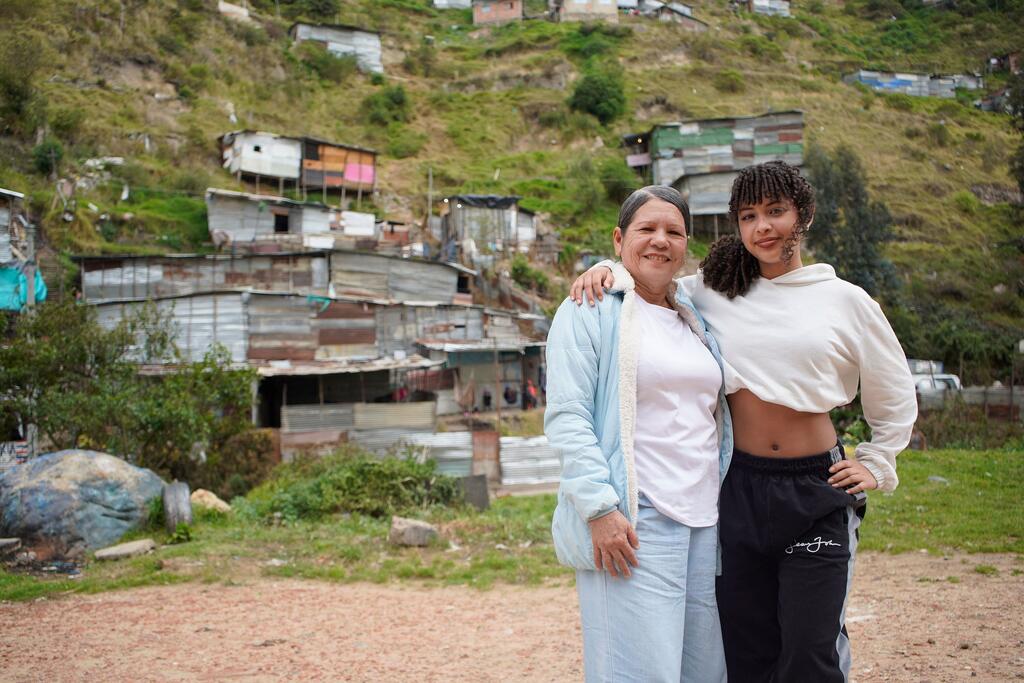 Dos mujeres latinoamericanas sonrientes se encuentran juntas en un barrio pobre colombiano, con casas improvisadas de cartón al fondo.