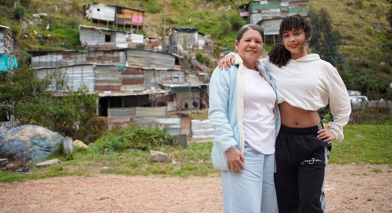 Dos mujeres latinoamericanas sonrientes se encuentran juntas en un barrio pobre colombiano, con casas improvisadas de cartón al fondo.