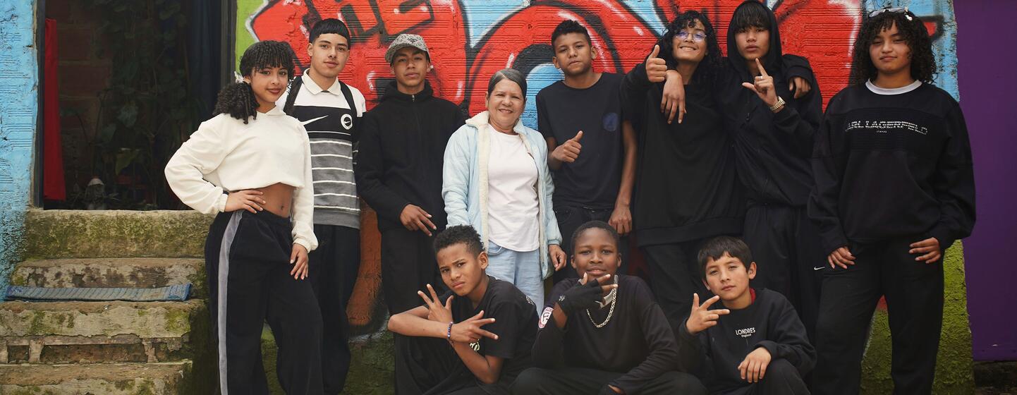 Un grupo de jóvenes colombianos y venezolanos posando juntos frente a un colorido muro de graffiti en el distrito Ciudad Bolívar de Bogotá, Colombia. La imagen captura la integración de la comunidad a través de la danza, destacando la vibrante vida cultural de este barrio urbano.