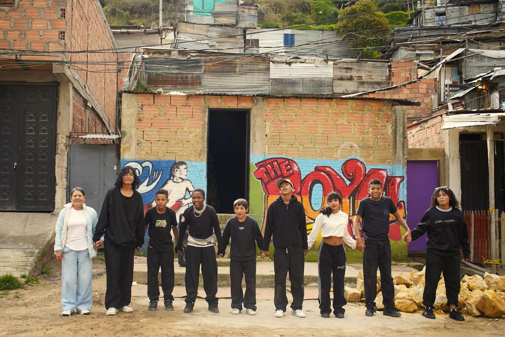 Un grupo de nueve colombianos y venezolanos, incluyendo adultos y niños, de pie en una fila tomándose de la mano frente a casas de ladrillo con graffiti en el distrito de Ciudad Bolívar de Bogotá, Colombia.