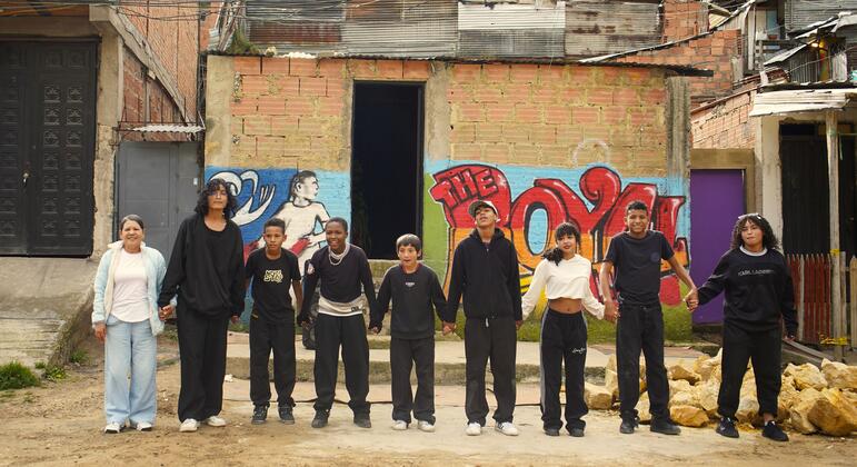 Un grupo de nueve colombianos y venezolanos, incluyendo adultos y niños, de pie en una fila tomándose de la mano frente a casas de ladrillo con graffiti en el distrito de Ciudad Bolívar de Bogotá, Colombia.