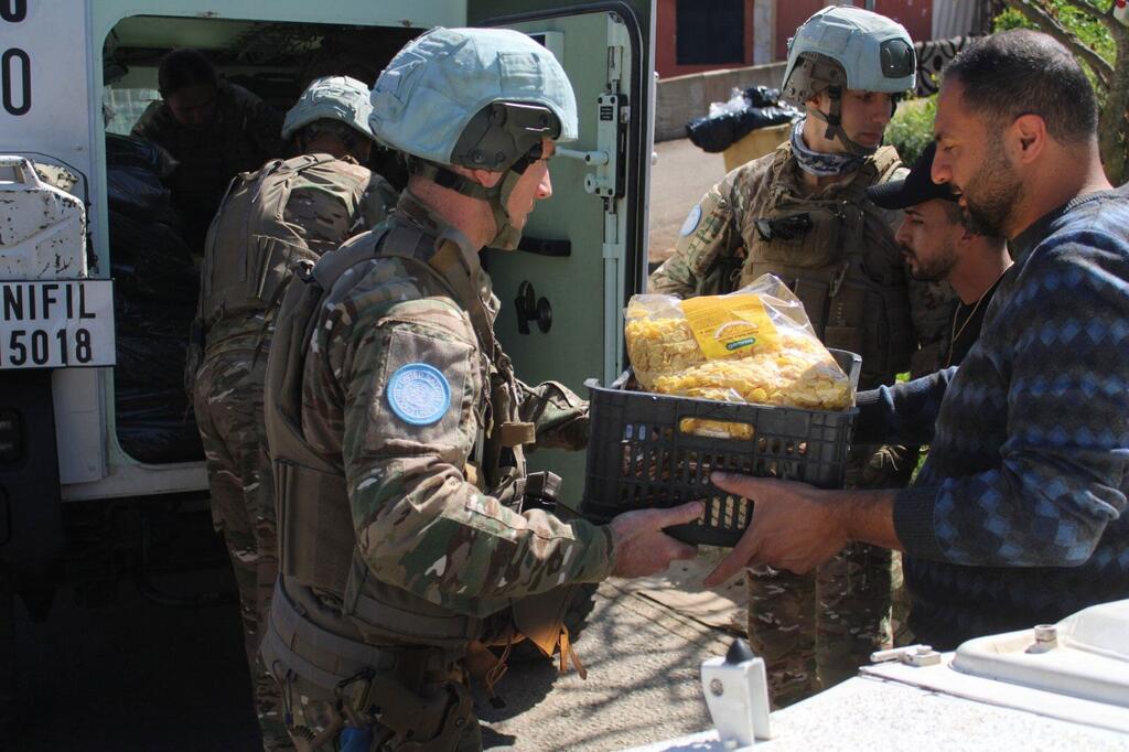 Wanajeshi wa kulinda amani wa UNIFIL wakiwa wamevaa mavazi ya kujificha na kofia za bluu wanagawanya sanduku la chakula kwa raia mmoja, huku askari mwingine akiwa amesimama kando ya gari la kijeshi nyuma.