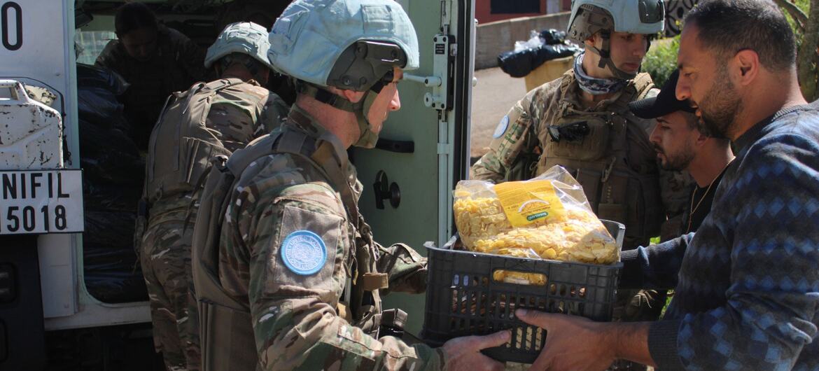 Fuerzas de paz de la UNIFIL con uniformes de camuflaje y cascos azules distribuyen una caja de alimentos a un civil, mientras que otro soldado se encuentra junto a un vehículo militar en el fondo.