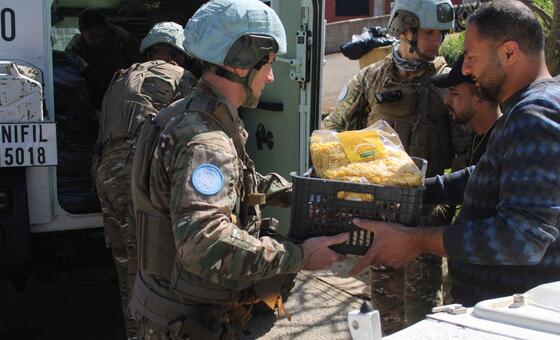 UNIFIL peacekeepers in camouflage uniforms and blue helmets distribute a crate of food to a civilian man, while another soldier stands by a military vehicle in the background.