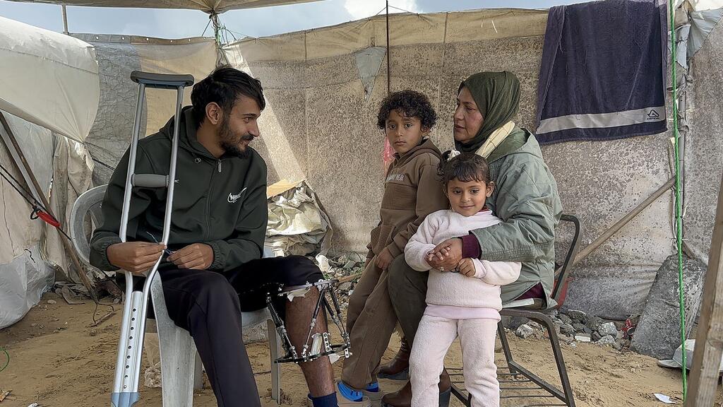Hajar Abed, miembro del personal de la UNRWA, se sienta con su familia en una tienda de campaña improvisada en Gaza, junto a ella se sienta un joven con un brazalete y muletas, mientras ella sostiene a una niña y un niño se sientan a su lado.