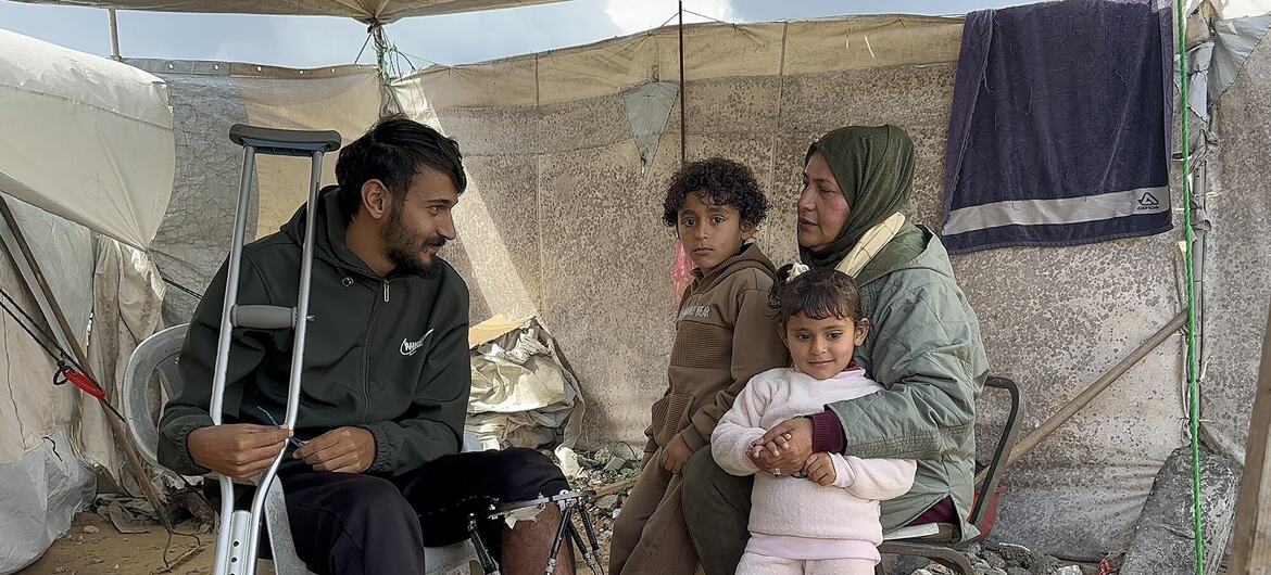 Hajar Abed, miembro del personal de la UNRWA, se sienta con su familia en una tienda de campaña improvisada en Gaza, junto a ella se sienta un joven con un brazalete y muletas, mientras ella sostiene a una niña y un niño se sientan a su lado.