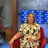 Uma jornalista com cabelos loiros e um vestido floral sorri sentada numa cadeira vermelha num estúdio de televisão com a marca ONU News visível nas telas atrás dela.