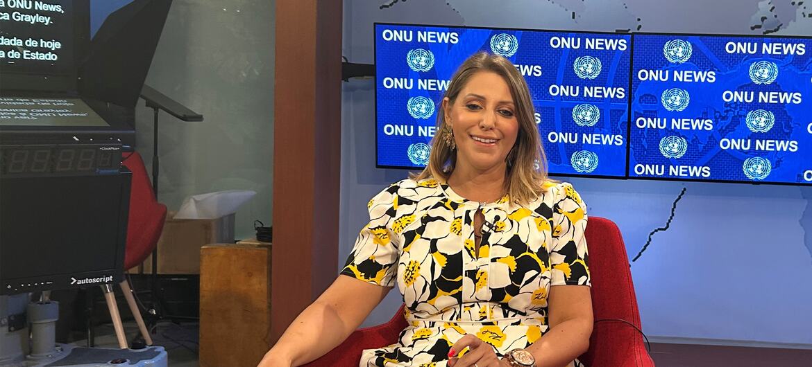 Uma jornalista com cabelos loiros e um vestido floral sorri sentada numa cadeira vermelha num estúdio de televisão com a marca ONU News visível nas telas atrás dela.