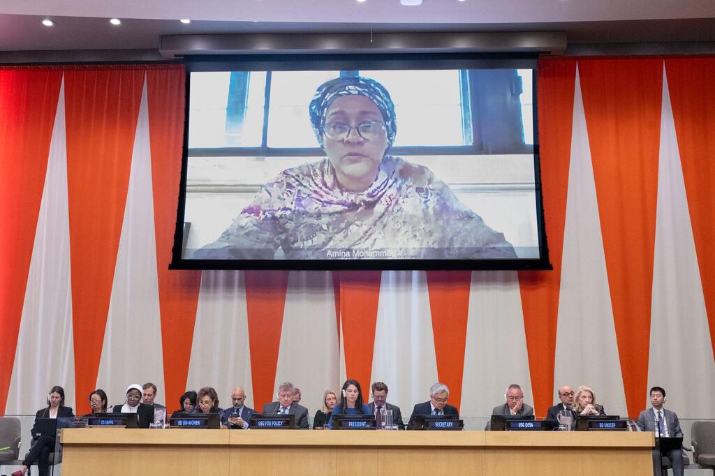 Deputy Secretary-General Amina Mohammed speaks via video link on a large screen at a UN General Assembly plenary meeting. Delegates are seated at a long table in the foreground.