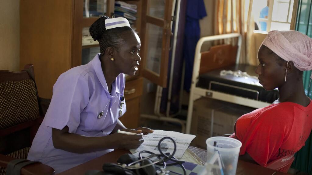 Muuguzi Mwafrika mwenye yunifomu ya zambarau anashauriana na mgonjwa wa kike aliyevaa kitambaa cha rangi ya waridi katika kliniki.