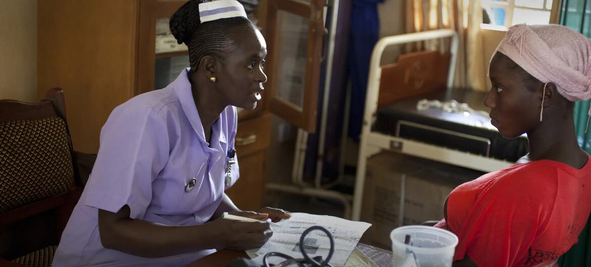 बैंगनी वर्दी में एक अफ्रीकी नर्स एक क्लिनिक में गुलाबी हेडस्क्रैप पहने हुए एक महिला रोगी से परामर्श करती है।