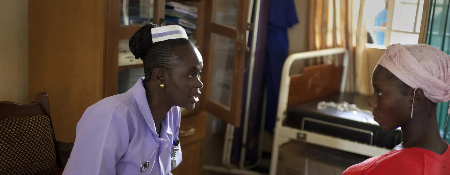 An African nurse in a purple uniform consults with a female patient wearing a pink headscarf in a clinic.
