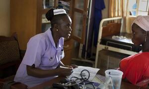 An African nurse in a purple uniform consults with a female patient wearing a pink headscarf in a clinic.