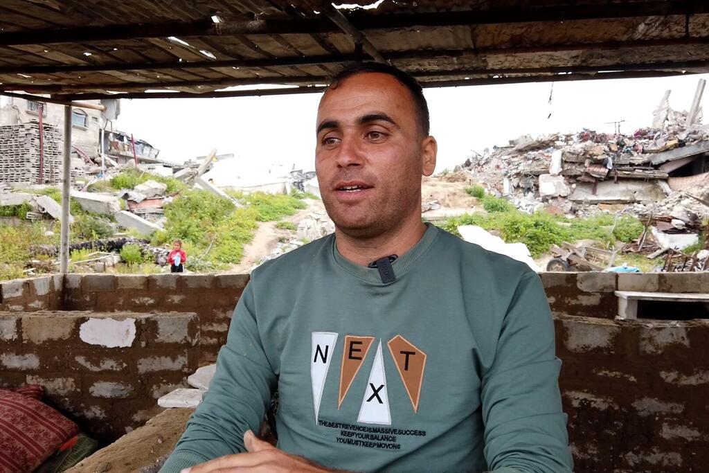 صورة لمحمد الجثبه يجلس في ملجأ مؤقت مبني من الأنقاض، مع مبان مدمرة مرئية في الخلفية في غزة.