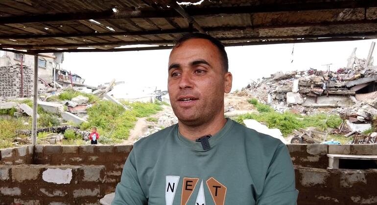 صورة لمحمد الجثبه يجلس في ملجأ مؤقت مبني من الأنقاض، مع مبان مدمرة مرئية في الخلفية في غزة.