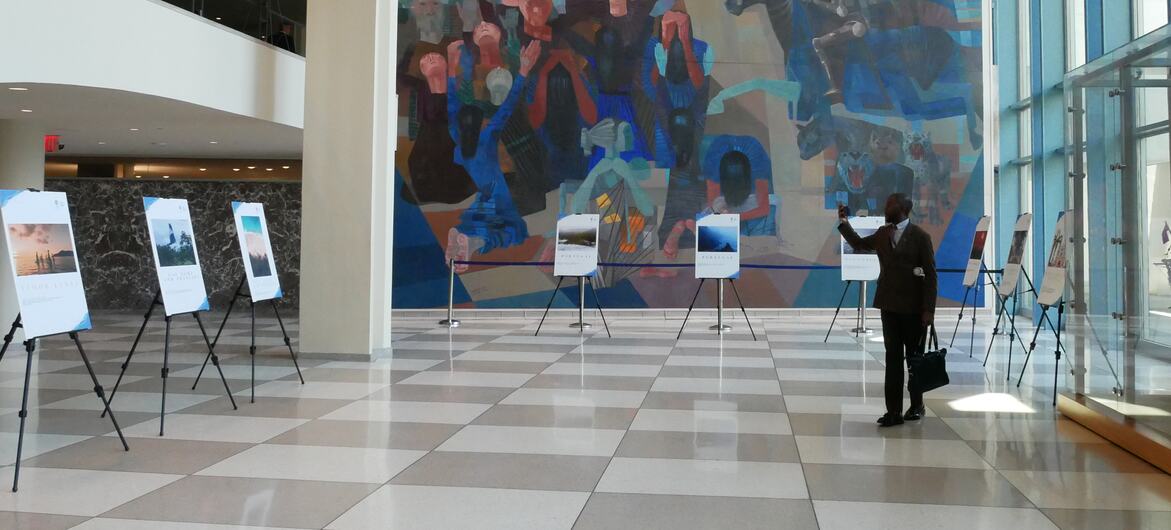 Um homem de terno tira uma foto de uma exposição de fotografias exibidas em cavaletes no saguão da sede das Nações Unidas, com um grande mural colorido ao fundo.
