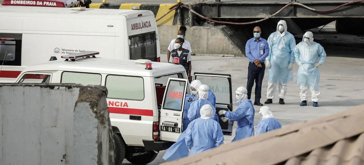 Medical personnel in blue protective suits stand near ambulances at a port during a Hantavirus evacuation coordinated by the WHO.