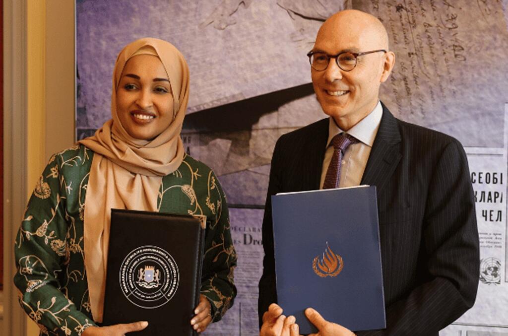 Kamishna Mkuu wa Umoja wa Mataifa wa Haki za Binadamu, Navi Pillay, anatabasamu huku akishikilia folda ya hati ya bluu kando ya mwanamke wa Somalia aliyevaa vazi la kijani na hijab ya rangi ya beji, akishikilia folda nyeusi yenye muhuri rasmi.