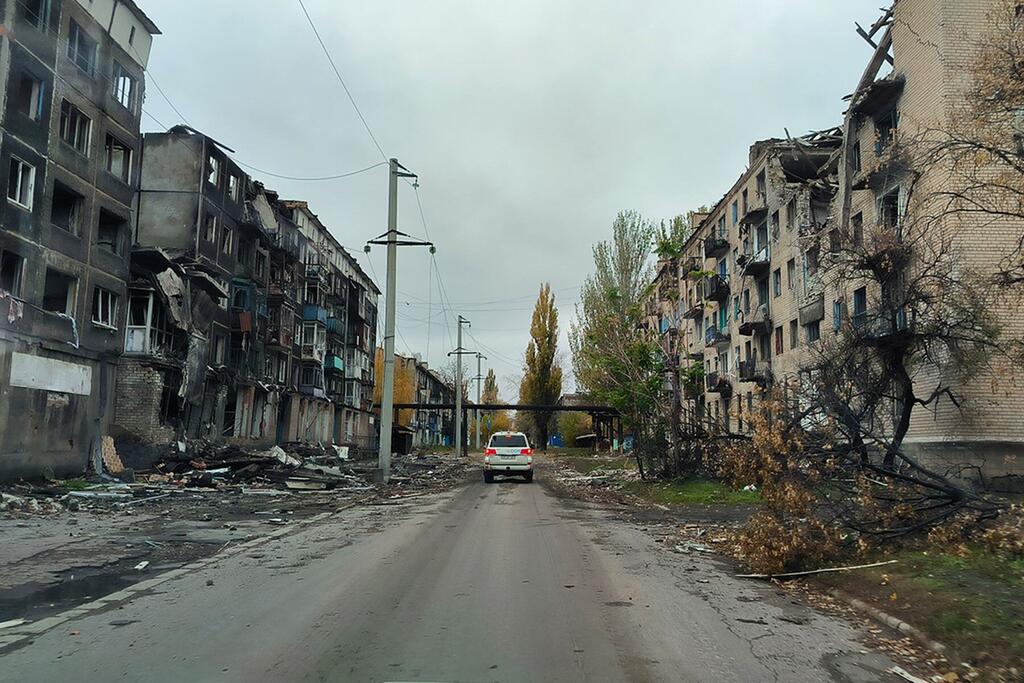 Un coche blanco desciende por una calle dañada en Ucrania, flanqueada por edificios de apartamentos fuertemente bombardeados con ventanas rotas y fachadas carbonizadas.