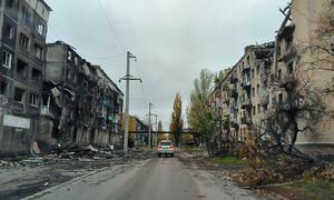 A white car drives down a damaged street in Ukraine, flanked by heavily bombed apartment buildings with broken windows and charred facades.