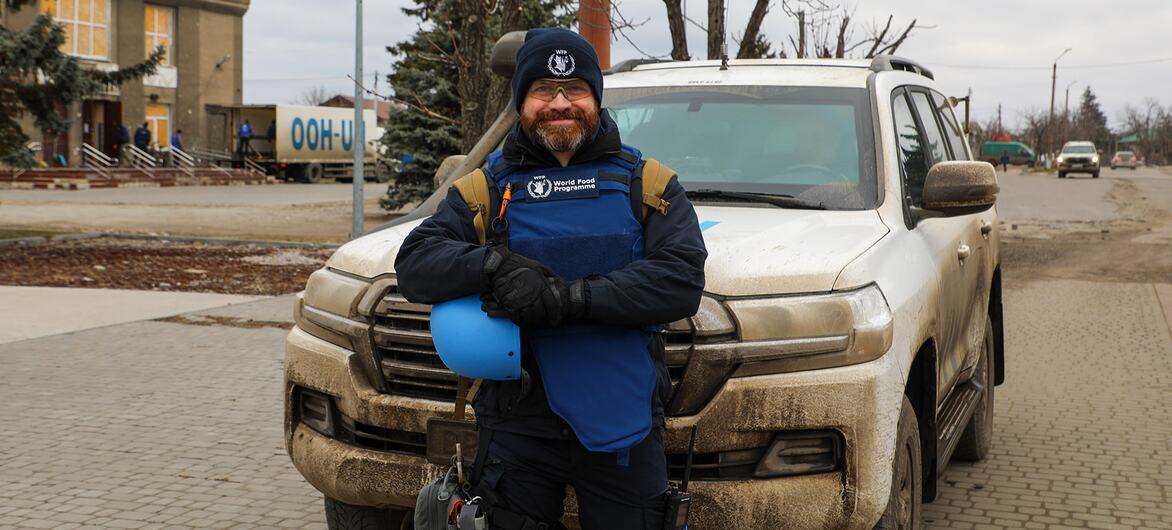 Работник Всемирной продовольственной программы стоит перед грязным белым внедорожником с синей шлемкой в украинском городе.