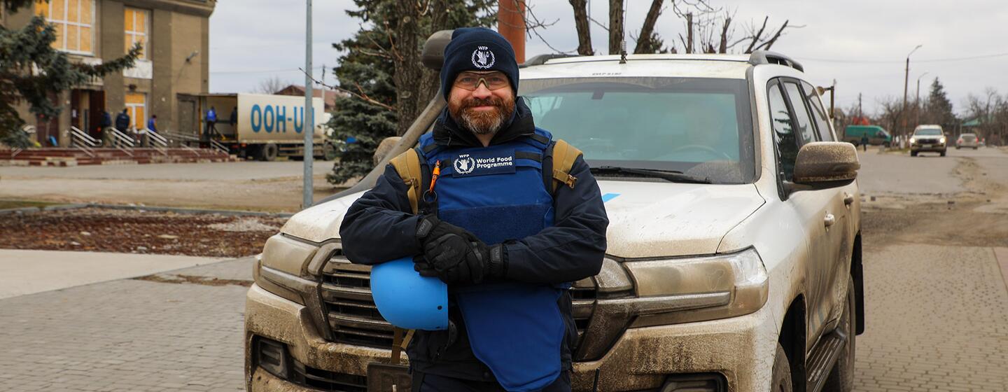 A World Food Programme worker stands in front of a muddy white SUV, holding a blue helmet, in a Ukrainian town.